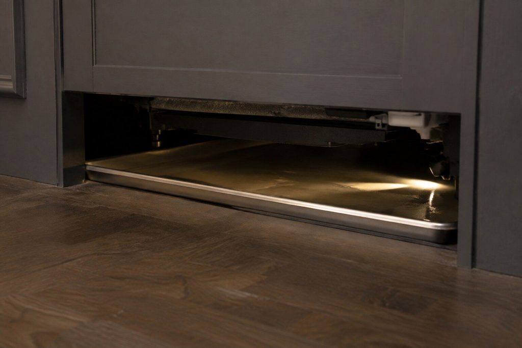 A shallow steel tray under a dishwasher seen low through the gap beneath a cabinet plinth, a thin film of water in it catching a torch beam.