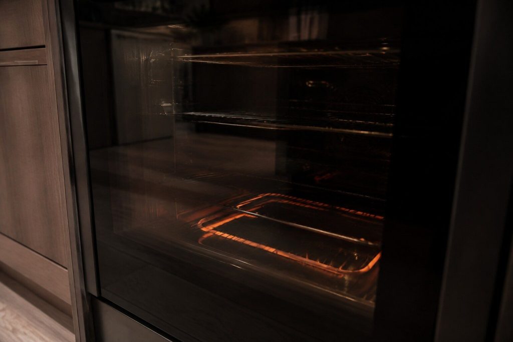 The lower element of an oven cavity glowing dull orange in an otherwise dark oven interior, seen through the door glass from slightly to one side.
