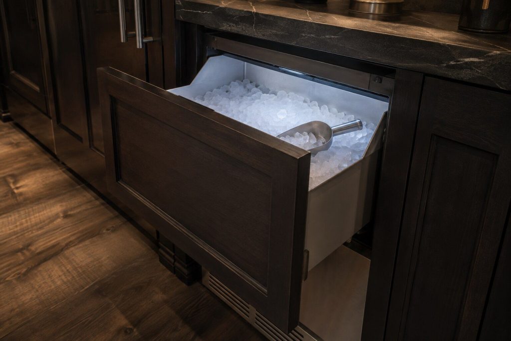 The open storage bin of a built-under ice machine heaped with small soft nugget ice, cool light falling in from the cabinet above and a metal scoop resting in the ice.