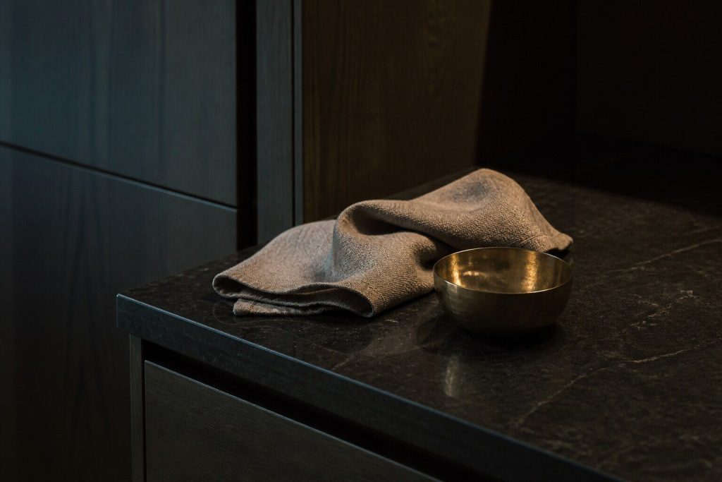 A quiet corner of a dark stone kitchen worktop in warm low light with a folded cloth and a small brass bowl on it, deep shadow behind.