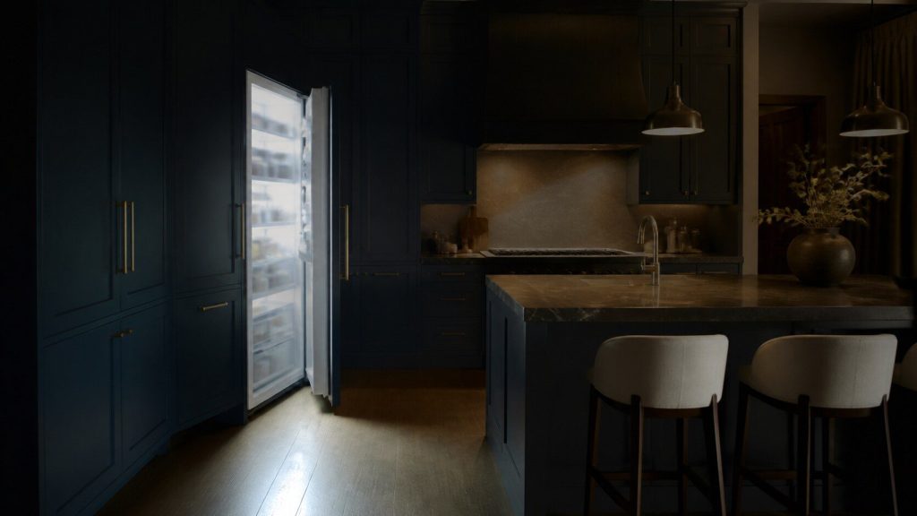 A wide dark kitchen at night with one tall integrated refrigerator door standing open, cold pale light spilling out of it across the floor and meeting the warm light of the room.