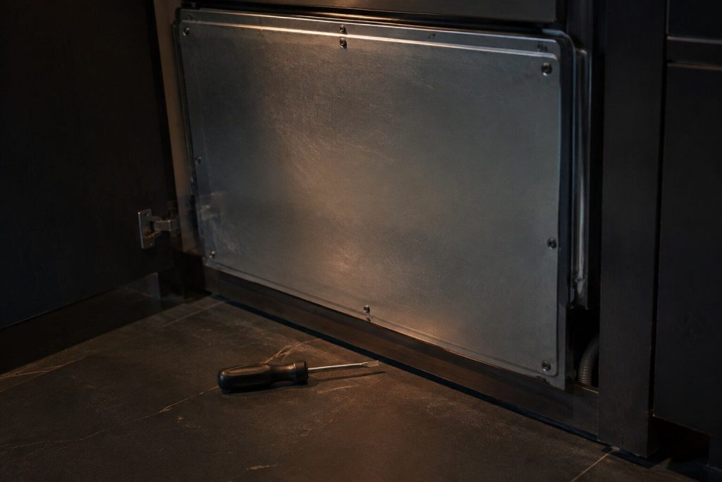 A rear service panel screwed shut on the back of a built-in appliance in a dark utility space, a single screwdriver laid down on the floor beside it and nobody touching it, one hard light raking across the metal.