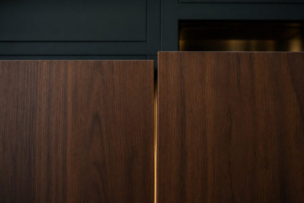 A tight detail of two tall cabinet doors meeting in a dark luxury kitchen, photographed square on, where the top of the left door sits a few millimeters below the top of the right one so the horizontal line between them is broken.