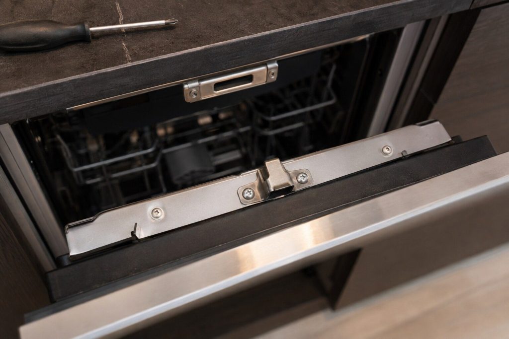 A close view of the latch tongue on the top edge of an open integrated dishwasher door and the striker plate above it in the tub frame, warm light, shallow depth of field.