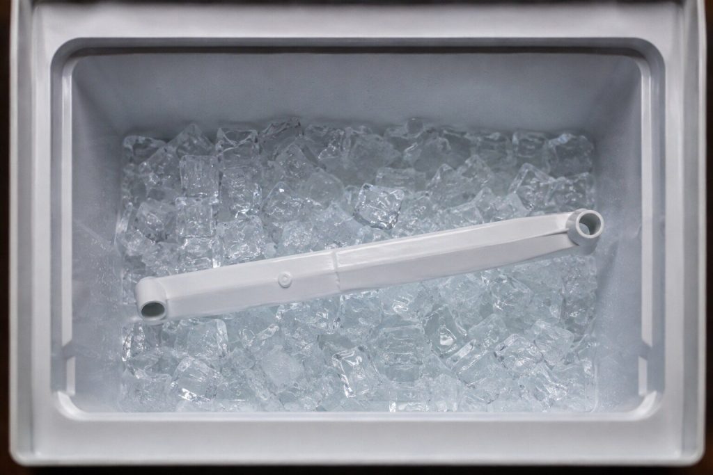 A moulded ice bin half full of clear cubes photographed from above in cold light, the harvest arm resting across the surface of the ice.