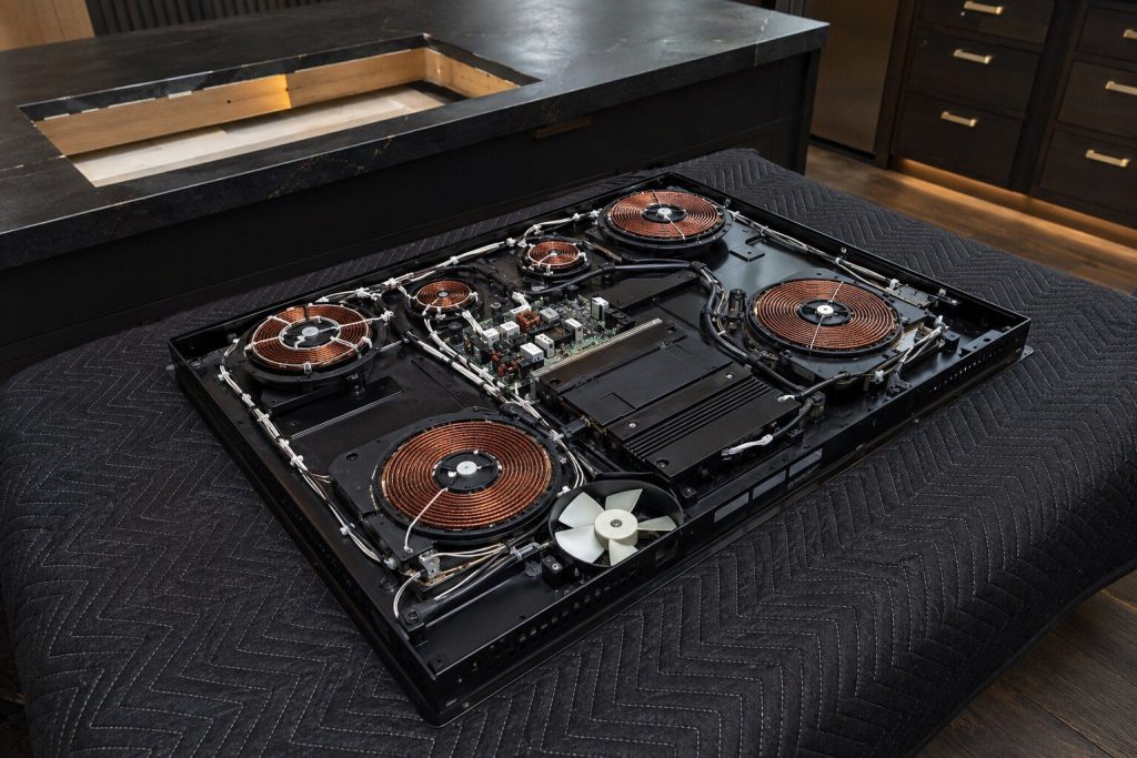 An induction cooktop lifted out of the worktop and turned face down on a padded blanket, the round copper induction coils and the cooling fan visible across its underside, cool workshop light.