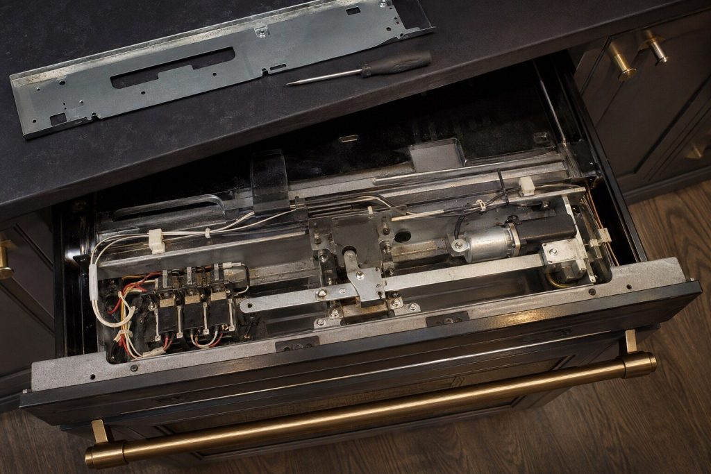 The motorized door latch assembly of a wall oven seen from above with the top trim removed — a small motor, a moving bar and a switch pack — warm torchlight on the metal.