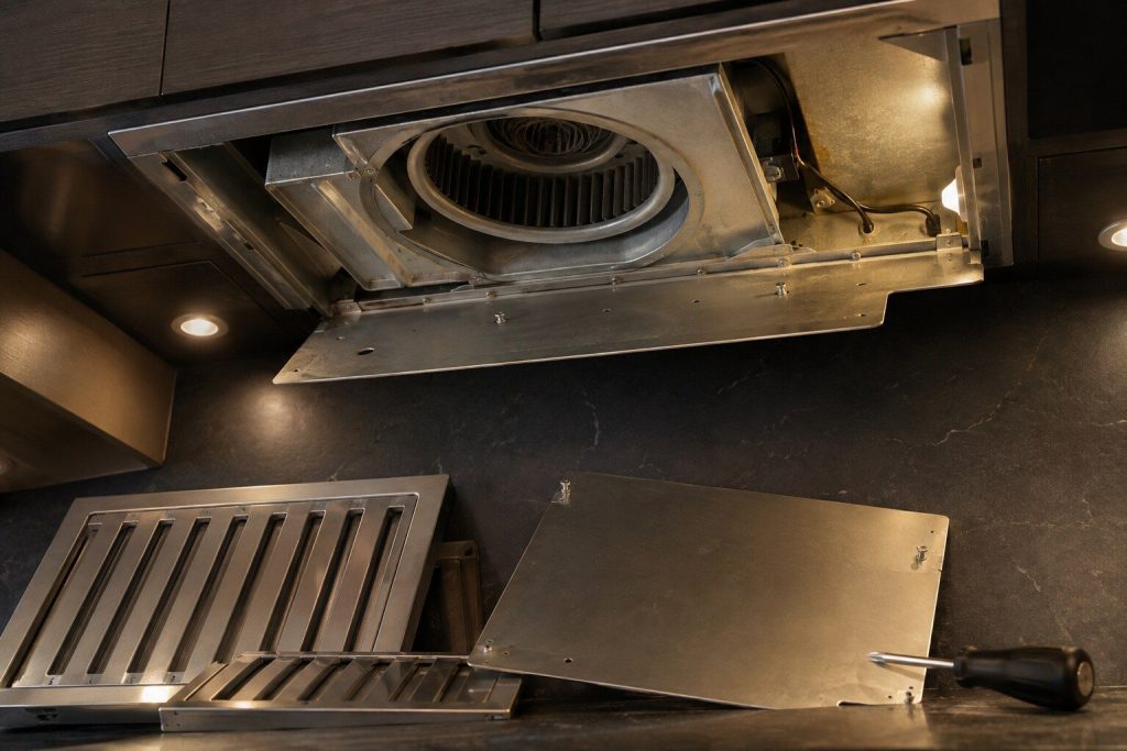 The inside of an extractor hood with the baffle filters removed, showing the blower housing and the fan wheel above, warm torchlight and a light film of grease on the metal.