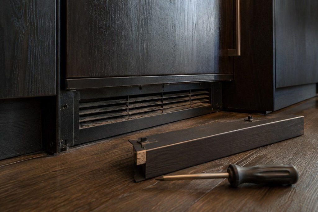 A low close view of the narrow ventilation grille at the bottom of an undercounter appliance door in a dark bar run, dust visible in the slots, warm light raking across it.