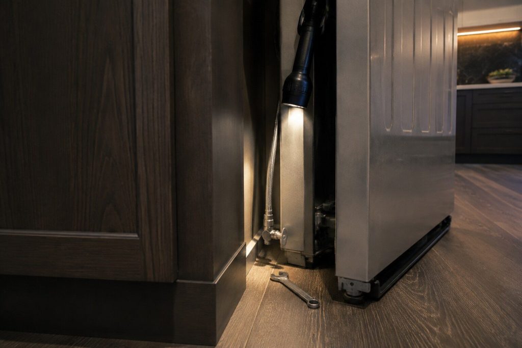 The braided water line and its small shut-off valve in the narrow gap behind a built-in refrigerator drawn forward from its opening, a spanner resting on the floor beside it, torch light down the gap.