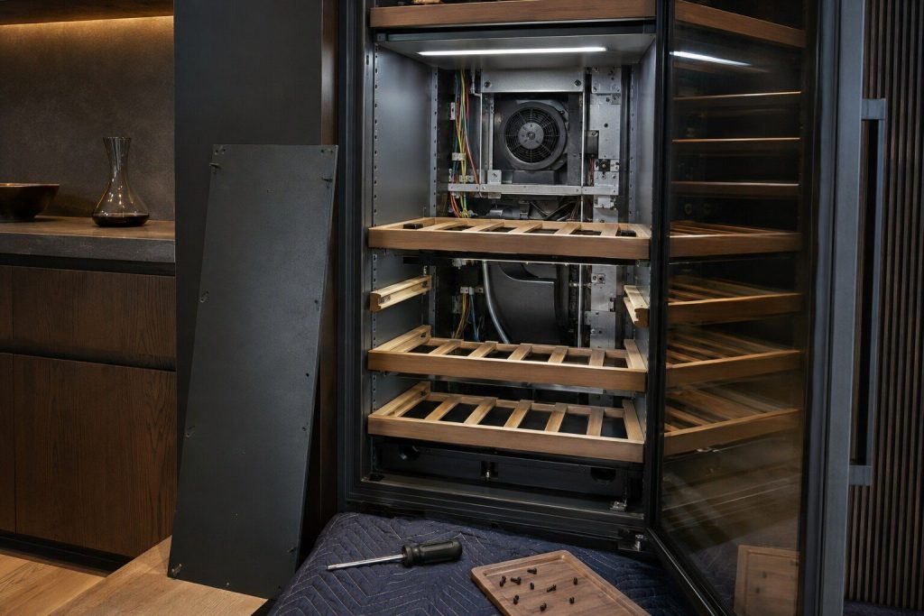 The circulation fan and rear duct of a built-in wine cabinet with the interior back panel unscrewed and leaning aside, the wooden shelves empty, cool light inside the cabinet.