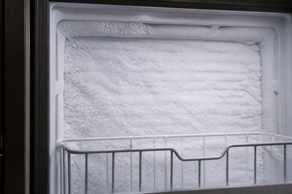 A thick even sheet of white frost across the moulded plastic back wall of a domestic freezer compartment, a wire basket edge in the foreground, photographed close in cold light so the texture of the ice reads clearly.