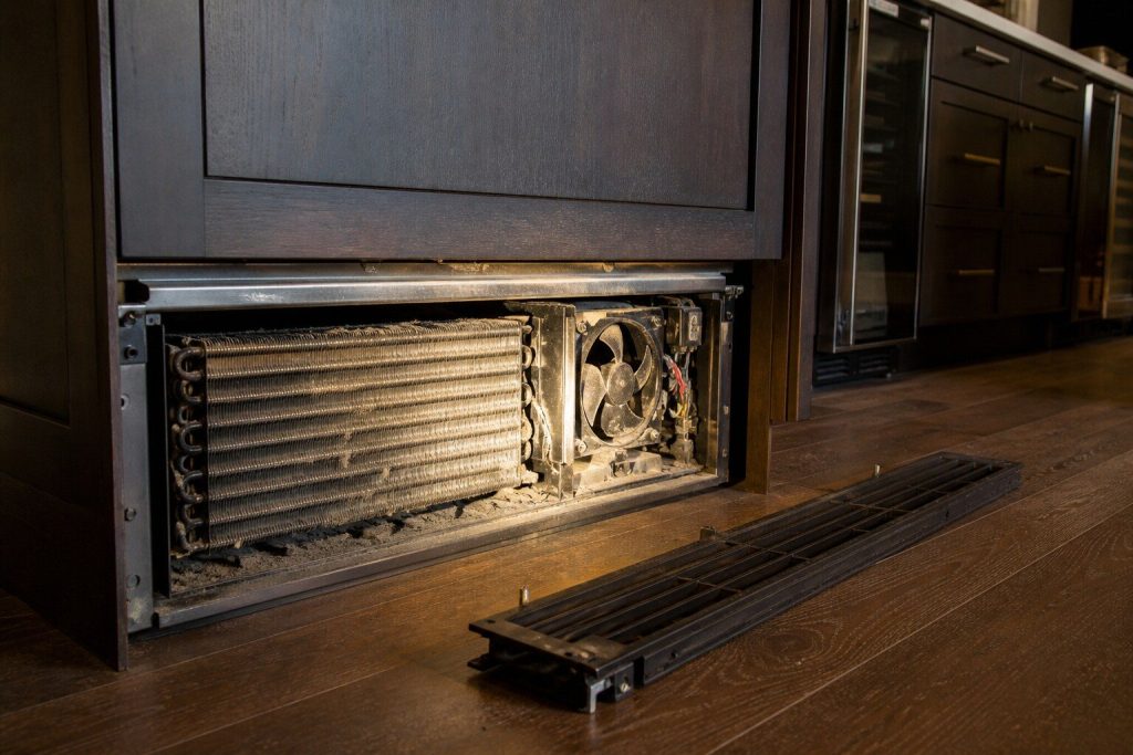 The lower grille of a tall built-in refrigerator removed and set aside, showing a dusty condenser coil and a fan in the machinery compartment behind it.