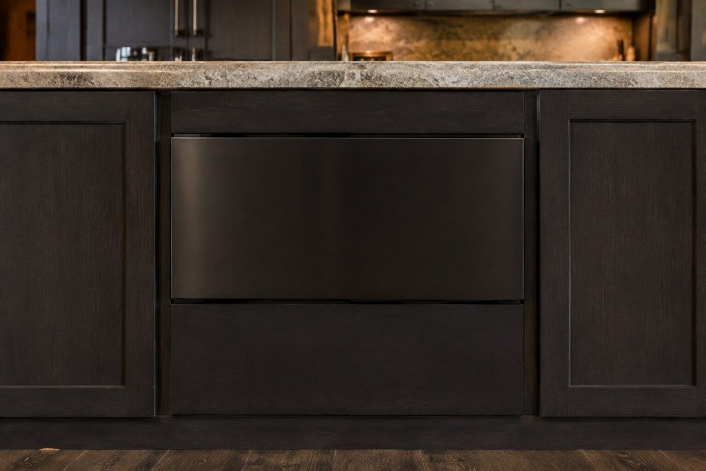 A microwave drawer front set into a dark kitchen island, closed, seen straight on with stone worktop above and cabinet doors either side.