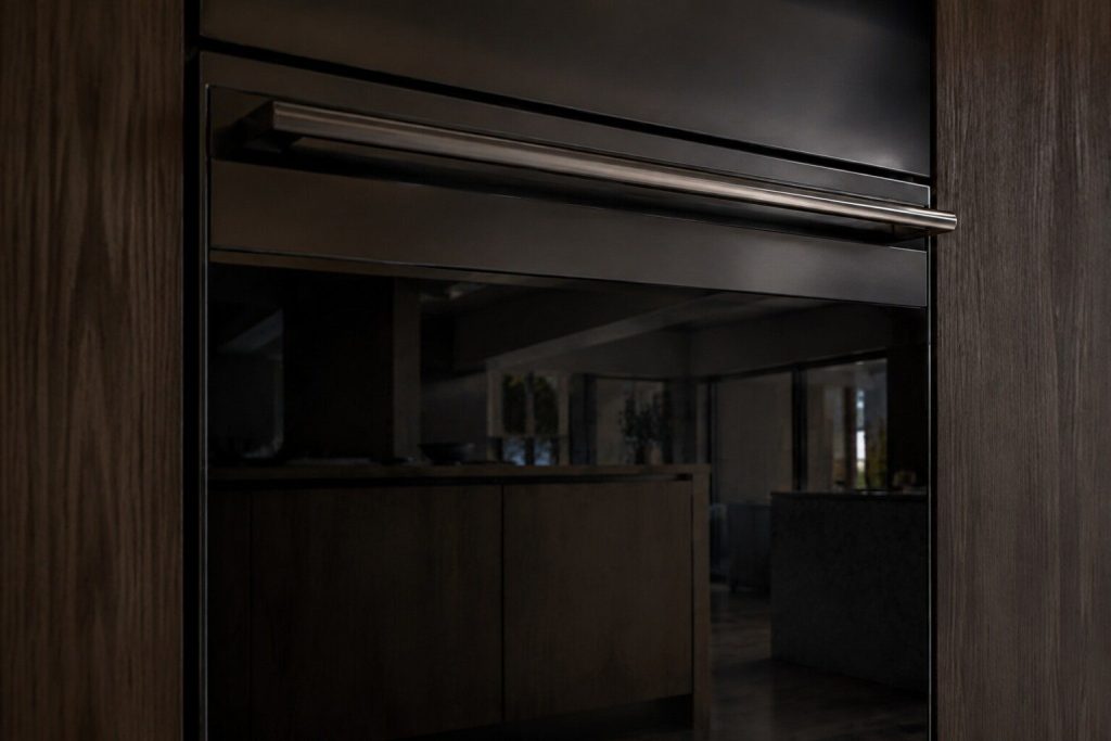The closed door of a built-in wall oven seen from a low angle in a dark kitchen, the handle and the top edge of the door in frame and the glass reflecting the room.