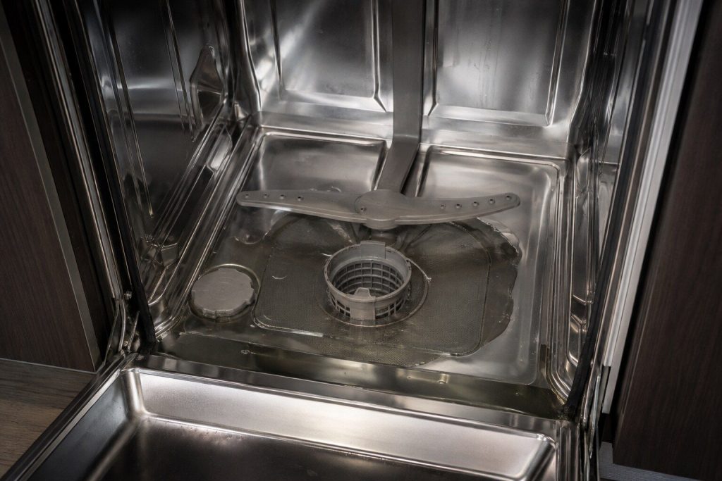 Standing water covering the floor of a stainless dishwasher tub around the cylindrical filter, the lower rack lifted out, seen from above in cool light.