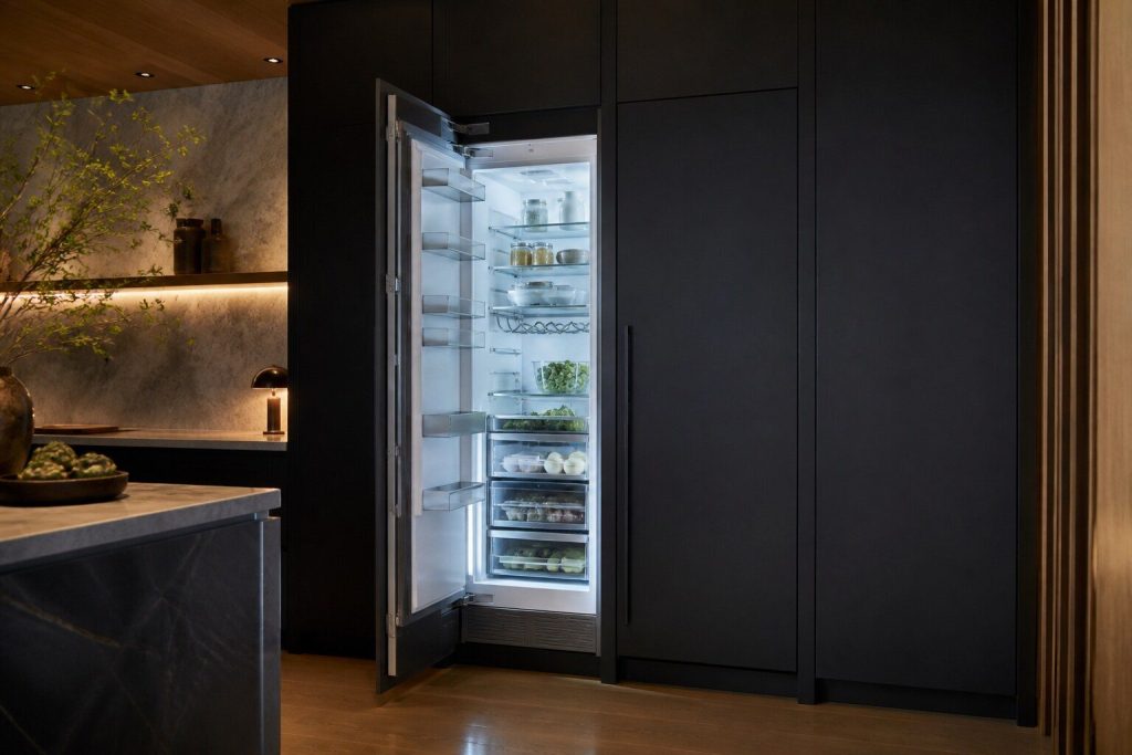 Two tall integrated column doors in a dark kitchen, one standing open showing lit shelves and the other closed and flush with the cabinetry beside it.