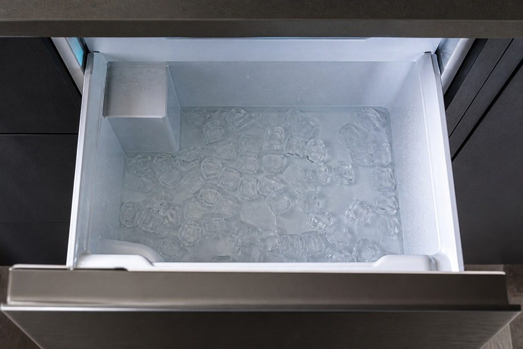The inside of an undercounter ice machine bin with a shallow layer of standing water under partly melted cubes, seen from above in cold light.