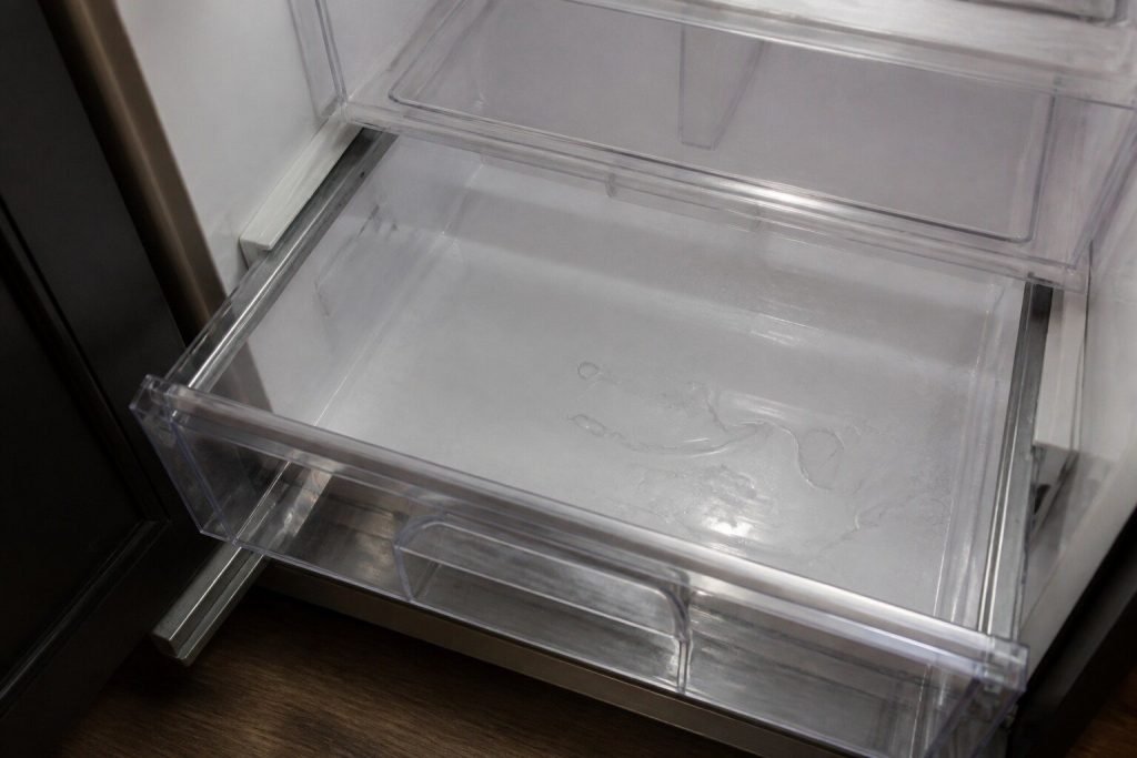 Water pooled across the floor of a refrigerator compartment beneath a clear crisper drawer that has been pulled forward, seen close and from above in cold interior light.
